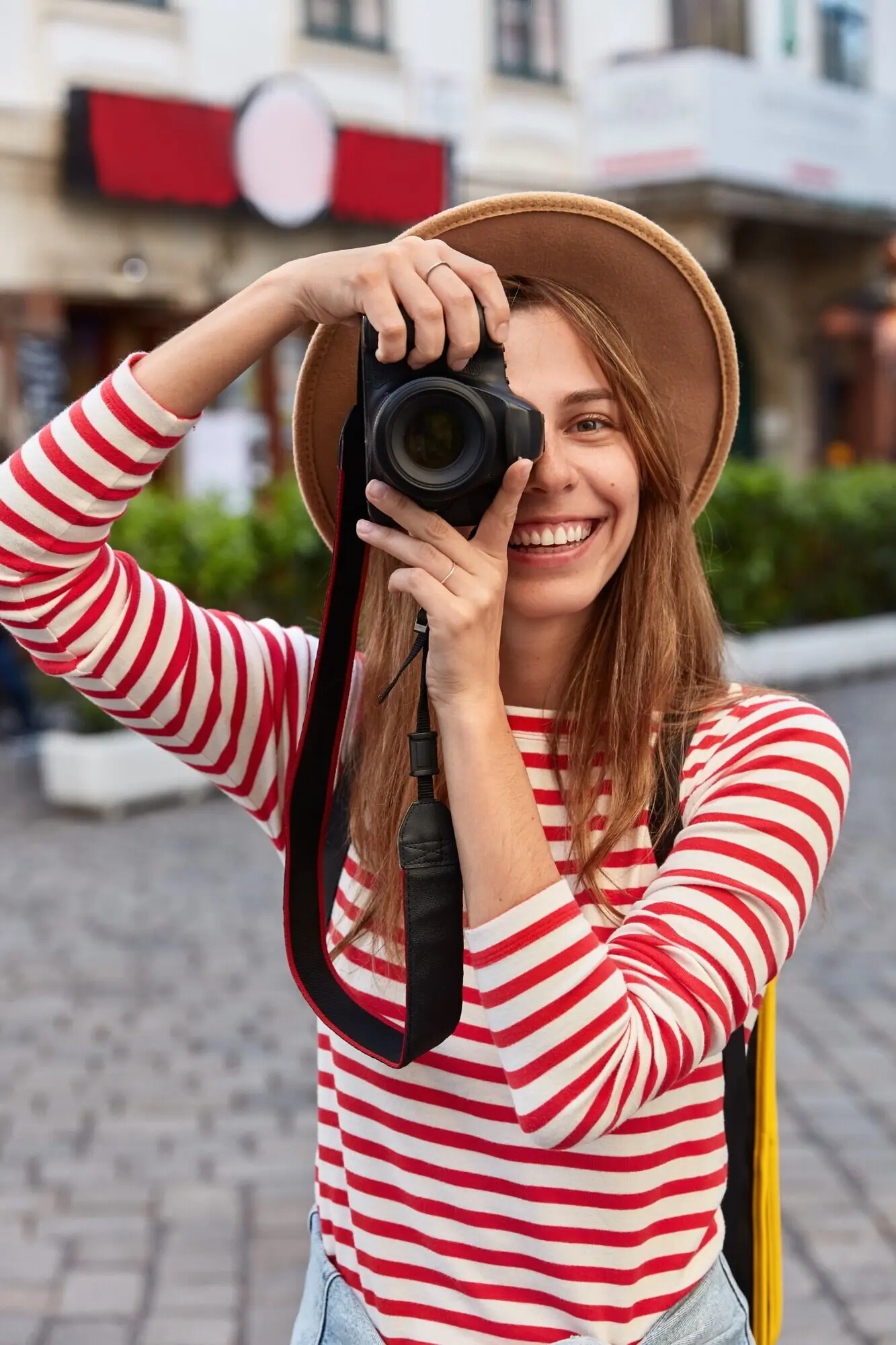 Fröhlicher Tourist macht mit einer Kamera professionelle Fotos, lächelt breit, richtet den Fokus auf ein schönes Wahrzeichen, schlendert durch das Stadtzentrum und trägt einen Hut.